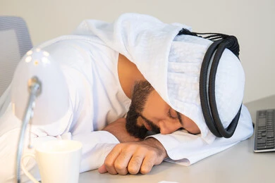 Gestures of drowsiness, fatigue and exhaustion, an Arab Gulf Emirati man wearing a white kandura and ghutra sitting behind a desk in the office taking a break, working in UAE companies, office profession and job, sleeping on the work desk