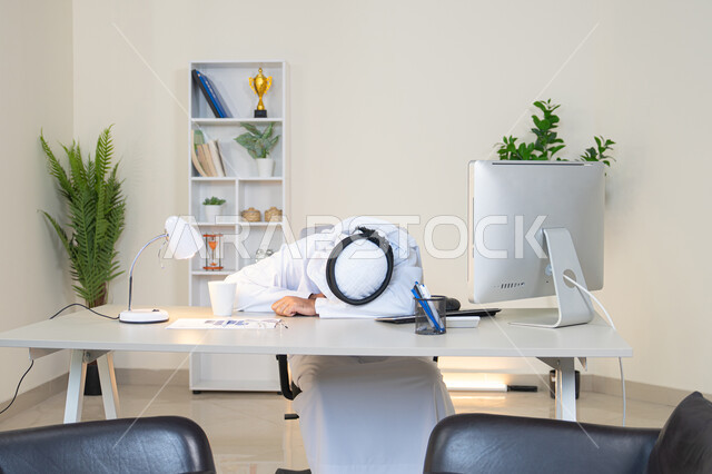النوم على طاولة العمل، ايماءات النعاس والتعب والإرهاق، رجل عربي خليجي اماراتي يرتدي الكندورة والغترة البيضاء يجلس خلف الطاولة في المكتب ويقوم بأخذ قسط من الراحة، العمل في شركات الامارات العربية المتحدة، مهنة ووظيفة مكتبية