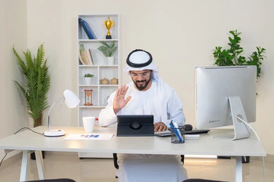 رفع كف اليد بايماءات القاء التحية والسلام، إجراء مكالمة فيديو، إنجاز مهام العمل، إدارة شؤون الأعمال وتنظيمها عبر الايباد، استخدام جهاز تقني حديث ومتطور، رجل عربي خليجي اماراتي يرتدي الكندورة والغترة البيضاء يجلس خلف الطاولة في المكتب ويستخدم الجهاز اللوحي