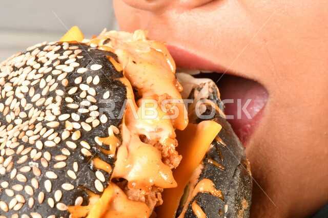 Burger sandwich with layers of meat and lettuce with melted cheese, a Saudi Arabian Gulf woman eating a burger, enjoying eating a burger, fast food, the harms of eating a lot of fast food