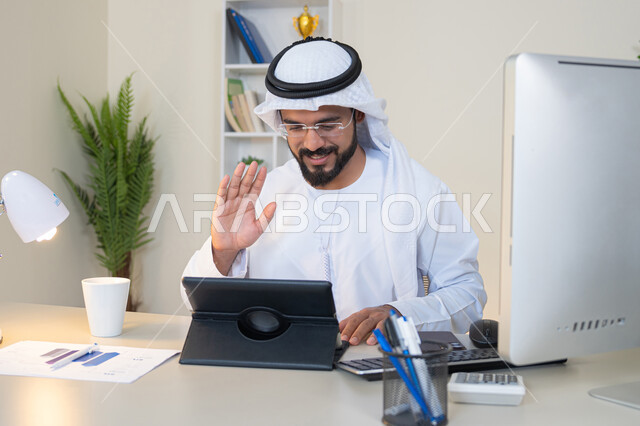 رفع كف اليد بايماءات القاء التحية والسلام، إجراء مكالمة فيديو، إنجاز مهام العمل، إدارة شؤون الأعمال وتنظيمها عبر الايباد، استخدام جهاز تقني حديث ومتطور، رجل عربي خليجي اماراتي يرتدي الكندورة والغترة البيضاء يجلس خلف الطاولة في المكتب ويستخدم الجهاز اللوحي