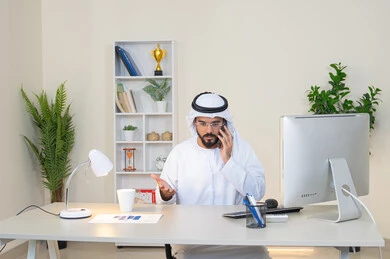 Sipping your favorite drink at the office, an Emirati Gulf Arab man wearing a white kandura and ghutra sitting behind a table holding a mobile phone and a cup of coffee next to him, working in a UAE company, an office job and profession, making a phone call, using a modern and advanced technical device