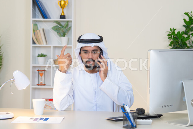 Making a phone call, using a modern and advanced technical device, drinking your favorite drink in the office, an Arab Gulf Emirati man wearing a white kandura and ghutra sitting behind a table holding a mobile phone and a cup of coffee next to him, working in a company in the United Arab Emirates, an office profession and job