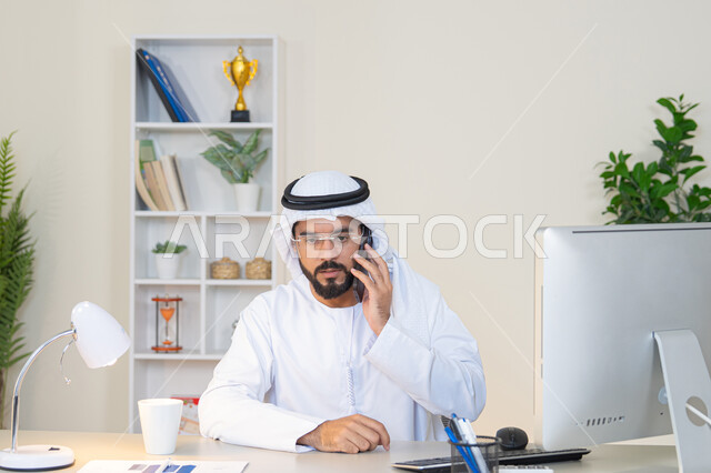 Making a phone call, using a modern and advanced technical device, drinking your favorite drink in the office, an Arab Gulf Emirati man wearing a white kandura and ghutra sitting behind a table holding a mobile phone and a cup of coffee next to him, working in a company in the United Arab Emirates, an office profession and job