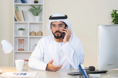 العمل في شركات الامارات العربية المتحدة، مهنة ووظيفة مكتبية، اجراء مكالمة هاتفية، استخدام جهاز تقني حديث ومتطور، احتساء المشروب المفضل في المكتب، رجل عربي خليجي اماراتي يرتدي الكندورة والغترة البيضاء يجلس خلف الطاولة يمسك بيده الهاتف المحمول وبجانبه كوب من القهوة