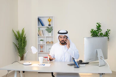 احتساء المشروب المفضل في المكتب، رجل عربي خليجي اماراتي يرتدي الكندورة والغترة البيضاء يجلس خلف الطاولة يمسك بيده الهاتف المحمول وبجانبه كوب من القهوة، العمل في شركات الامارات العربية المتحدة، مهنة ووظيفة مكتبية، اجراء مكالمة هاتفية، استخدام جهاز تقني حديث ومتطور