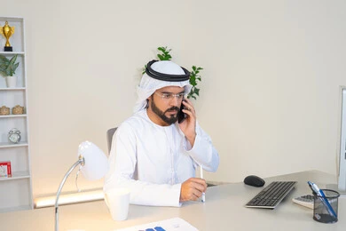 اجراء مكالمة هاتفية، استخدام جهاز تقني حديث ومتطور، احتساء المشروب المفضل في المكتب، رجل عربي خليجي اماراتي يرتدي الكندورة والغترة البيضاء يجلس خلف الطاولة يمسك بيده الهاتف المحمول وبجانبه كوب من القهوة، العمل في شركات الامارات العربية المتحدة، مهنة ووظيفة مكتبية