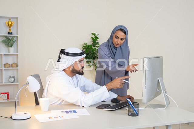 Pointing the index finger at the computer screen, exchanging experiences and information to achieve goals, team spirit and teamwork, understanding and cooperation between colleagues and developing strategic plans, holding a meeting at the company headquarters, two Arab Gulf Emirati employees learning work methods and professional development