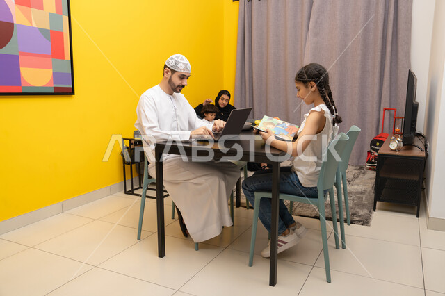 Doing business online from home, using modern and advanced technical devices, an Arab Gulf Emirati man next to his daughter using a laptop, a side view of an Arab Gulf Emirati family sitting in the living room