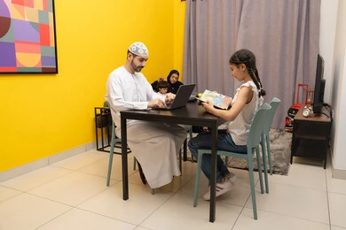 Doing business online from home, using modern and advanced technical devices, an Arab Gulf Emirati man next to his daughter using a laptop, a side view of an Arab Gulf Emirati family sitting in the living room