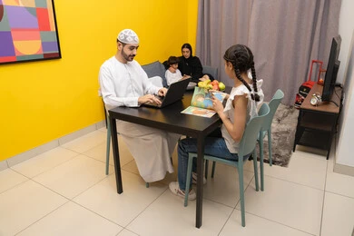 الاشراف على المخططات وتحليل اداء العمل، جلسه عمل عن بعد، استخدام الأجهزة التقنية الحديثة والمتطورة ، رجل عربي خليجي اماراتي  بجانب ابنته يقوم بأستخدام الحاسوب المحمول، صورة من الجانب لعائله عربيه خليجيه اماراتية تجلس داخل غرفه المعيشة في المنزل