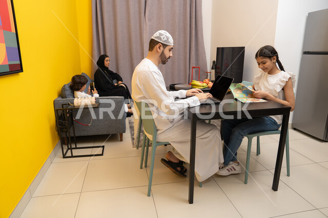 العمل عن بعد في المنزل برفقة الأبناء، استخدام الأجهزة التقنية الحديثة والمتطورة ، رجل عربي خليجي اماراتي  بجانب ابنته يقوم بأستخدام الحاسوب المحمول، صورة من الجانب لعائله عربيه خليجيه اماراتية تجلس داخل غرفه المعيشة في المنزل
