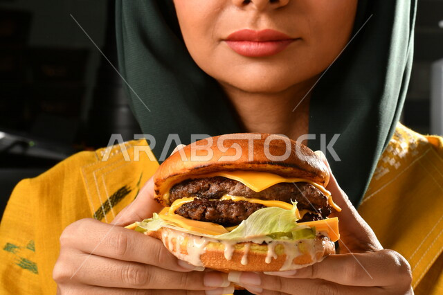 Burger sandwich with layers of meat and lettuce with melted cheese, a Saudi Arabian Gulf woman eating a burger, enjoying eating a burger, fast food, the harms of eating a lot of fast food
