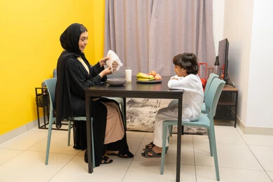 بناء علاقة متينة مع الأبناء، امراة عربيه خليجيه اماراتية ترتدي العباءه السوداء تجلس على الطاوله برفقه ابنها و تقوم بسكب الكورن فليكس و تحضيره ، طفل عربي خليجي يقوم بالنظر الى والدته بايماءات السعاده و السرور
