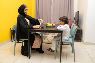 أجواء عائلية مليئة بالحب، امراة عربيه خليجيه اماراتية ترتدي العباءه السوداء تجلس على الطاوله برفقه ابنها و تقوم بسكب اللبن و تحضير الكورن فليكس ، طفل عربي خليجي يقوم بالنظر الى والدته بايماءات السعاده و السرور