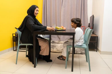 مفهوم الامومة والتربية بالعطف و الحنان، امراة عربيه خليجيه اماراتية ترتدي العباءه السوداء تجلس على الطاوله برفقه ابنها و تقوم بتقديم الطبق ، طفل عربي خليجي يقوم بالنظر الى والدته بايماءات السعاده و السرور