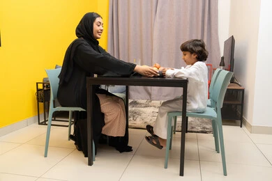 تقديم الرعاية والاهتمام للأبناء ، مفهوم التربية بالحب والحنان، امراة عربيه خليجيه اماراتية ترتدي العباءه السوداء تجلس على الطاوله برفقه ابنها و تقوم باطعامه ، طفل عربي خليجي يقوم بتناول طبق من الكورن فليكس ، ايماءات السعاده و السرور