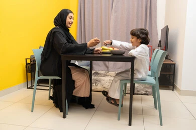 Caring for and supporting children, the concept of raising children with love and tenderness, an Arab Gulf Emirati woman wearing a black abaya sitting at the table with her son and feeding him, an Arab Gulf child eating a plate of cornflakes, gestures of happiness and pleasure