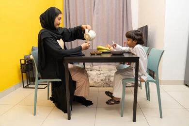 Spending quality time with children, enjoying drinks, the concept of raising children with love and tenderness, an Emirati Gulf Arab woman wearing a black abaya sitting at the table with her son and pouring a cup of tea, an Arab Gulf child eating a plate of cornflakes, gestures of happiness and pleasure