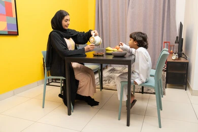 الاستمتاع بتناول المشروبات ، مفهوم التربية بالحب والحنان، امراة عربيه خليجيه اماراتية ترتدي العباءه السوداء تجلس على الطاوله برفقه ابنها و تقوم بصب كوب من الشاي ، طفل عربي خليجي يقوم بتناول طبق من الكورن فليكس ، ايماءات السعاده و السرور