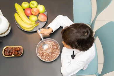 تناول الفواكه الصحية ، تصوير من الاعلي لطفل عربي خليجي اماراتي يرتدي الزي التقليدي و يجلس على الطاوله يقوم بتناول طبق من الكورن فليكس ، الوجبات السريعة والشهية، المنتجات الغذائية المهمة