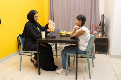 تحضير وتجهيز المأكولات والوجبات السريعة، وجبة طعام صحية، امراه عربية خليجية اماراتية مبتسمة محجبة ترتدي العباءة تجلس على الطاوله تقوم بسكب الكورن الفليكس و تحضير السناك برفقه ابنتها