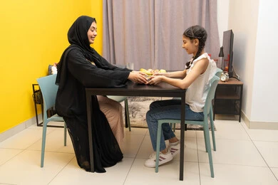 A family atmosphere full of love, the concept of motherhood and caring for children, a smiling veiled Emirati Gulf Arab woman wearing an abaya sitting at the table with her daughter with gestures of happiness and pleasure, a Gulf girl eating a plate of healthy snacks