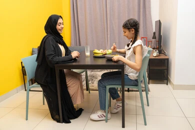مفهوم الامومه والاهتمام بالأولاد، امراه عربية خليجية اماراتية مبتسمة محجبة ترتدي العباءة تجلس على الطاوله برفقه ابنتها بايماءات السعاده و السرور، بنت خليجيه تقوم بتناول طبق من السناك الصحي