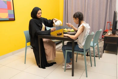 متعة لمة الاهل على السفرة، امراه عربية خليجية اماراتية مبتسمة محجبة ترتدي العباءة تجلس على الطاوله برفقه ابنتها و تقوم بصب المشروب المفضل، الاستمتاع بالمشروبات الساخنه، بنت خليجيه تقوم بتناول طبق من السناك الصحي