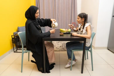 اجتماع العائلة على طاولة الطعام، امراه عربية خليجية اماراتية مبتسمة محجبة ترتدي العباءة تجلس على الطاوله برفقه ابنتها و تقوم بصب المشروب المفضل، الاستمتاع بالمشروبات الساخنه، بنت خليجيه تقوم بتناول طبق من السناك الصحي