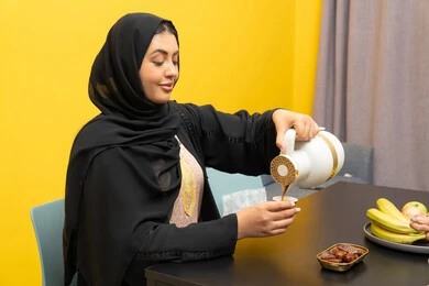 الشعور بالدفء والراحة، امراه عربية خليجية اماراتية مبتسمة محجبة ترتدي العباءة تجلس على كرسي تحمل تقوم بصب فنجان من الشاي بايماءات السرور، الاستمتاع بالمشروبات الساخنه