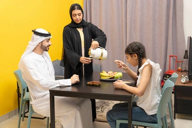 Sharing breakfast with family, eating a healthy meal, family breakfast, an Emirati Gulf Arab woman wearing a black abaya pouring tea for her husband, kindness and tenderness between family members, a Gulf girl eating cornflakes with milk
