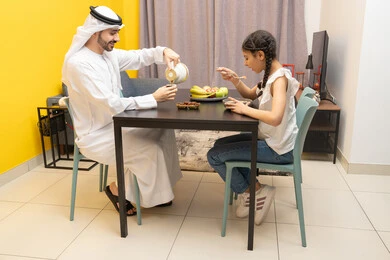Happy family atmosphere, eating a healthy meal, family breakfast, an Emirati Gulf Arab father sitting at the table pouring a cup of coffee with his daughter, affection and tenderness between family members, a Gulf girl eating cornflakes with milk