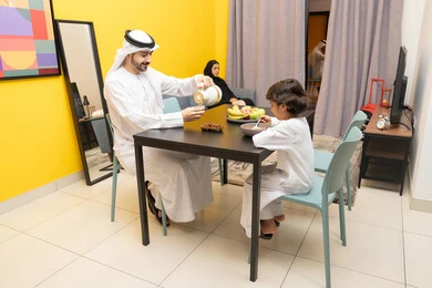 Fresh healthy fruits, traditional breakfast dishes, healthy meals and foods, an Emirati Gulf Arab father wearing a kandura and ghutra sitting at the table inside the living room with his son and pouring a cup of coffee, gestures of happiness and pleasure, a Gulf mother sitting on the couch watching TV