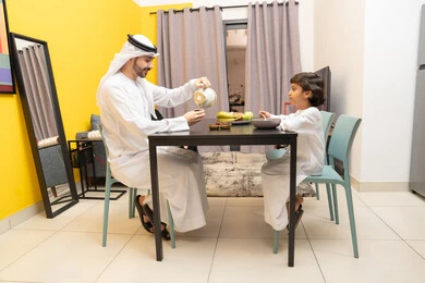 أطباق إفطار تقليدية، وجبات ومأكولات صحية، الفواكه الصحية الطازجة، اب عربي خليجي اماراتي يرتدي الكندورة و الغتره يجلس على الطاوله داخل عرفة المعيشة برفقه ابنه ، ايماءات السعاده و السرور ،طفل خليجي يقوم بتناول الطعام