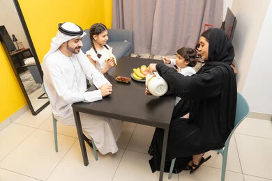 وجبات ومأكولات صحية، أطباق إفطار تقليدية، الفواكه الصحية الطازجة، عائله عربية خليحجيه اماراتية ترتدي الزي التقليدي تجلس على الطاوله داخل عرفة المعيشة ، ايماءات السعاده و السرور ، امراه خليجيه تقوم بصب كوب من القهوه