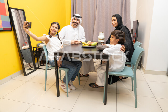 التقاط صور السيلفي عبر الجوال للذكريات العائلية الودية، مفهوم الترابط و التفاهم الأسري،عائله عربيه خليجيه اماراتية ترتدي الزي التقليدي تجلس على الطاوله داخل غرفه المعيشه ، طفله عربيه تمسك الهاتف المحمول بيدها بايماءات السعادة و السرور