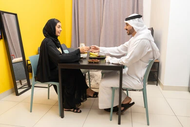 مفهوم الترابط و التفاهم الأسري، وجبة الفطور الصباحية، امراه عربيه خليجيه اماراتية ترتدي العباءة السوداء تجلس على الطاوله فى غرفه المعيشة برفقه زوجها تقوم باعطاء زوجها كوب من القهوه