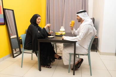 أجواء اسرية سعيدة في المنزل، وجبة الفطور الصباحية، امراه عربيه خليجيه اماراتية ترتدي العباءة السوداء تجلس على الطاوله فى غرفه المعيشة برفقه زوجها يقومان بتناول الطعام