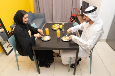 اجتماع الأسرة على السفرة، وجبة الفطور الصباحية، تدعيم أواصر المحبه والترابط الأسري، امراه عربيه خليجيه اماراتية ترتدي العباءة السوداء تجلس على الطاوله فى غرفه المعيشة برفقه زوجها يقومان بتناول الطعام
