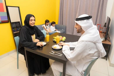 Having a good time, strengthening the bonds of love and family cohesion, an Arab Gulf Emirati family sitting in the living room inside the house, an Arab Gulf Emirati man wearing a kandura and ghutra sitting at the dining table with his wife eating breakfast, two Gulf children sitting on the couch watching TV