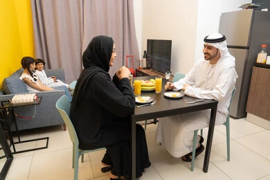 The joy of spending time with family, strengthening the bonds of love and family cohesion, an Arab Gulf Emirati family sitting in the living room inside the house, an Arab Gulf Emirati man wearing a kandura and ghutra sitting at the dining table with his wife eating breakfast, two Gulf children sitting on the sofa