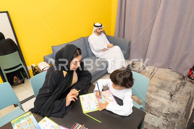 تنمية المهارات الفنية، مفهوم فنون الرسم عند الأطفال ، قضاء اوقات عائلية ممتعة في المنزل، ، ام عربيه خليجيه اماراتية ترتدي العباءة السوداء تجلس على الطاوله فى غرفه المعيشة تقوم بتعليم ومشاركة  ابنها بالتلوين، حضانة أطفال ، التطوير والإبداع