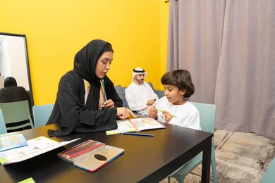 The concept of children's art, spending fun family time at home, an Emirati Gulf Arab mother wearing a black abaya sitting at the table in the living room teaching and participating with her son in coloring, developing artistic skills, nursery, development and creativity