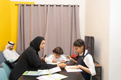 التعلم والابداع وتنمية المهارات، قضاء اوقات عائلية ممتعة في المنزل، لعبة تلوين الرسومات،عائله عربيه خليجيه اماراتية ترتدي الزي التقليدي يجلسون في غرفه المعيشه مع ابنائهم بأيماءات السعادة و السرور ، ام عربيه خليجيه تقوم بتعليم ومشاركة اولادها بالتلوين