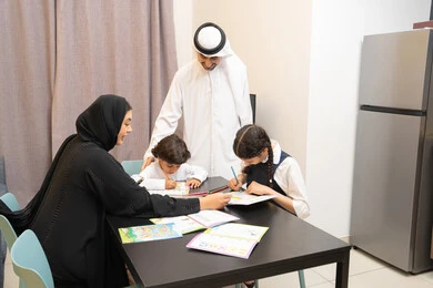 Educational activities and capacity building, spending fun family time at home, coloring game, an Emirati Gulf Arab family wearing traditional dress sitting in the living room with their children with gestures of happiness and pleasure, an Arab Gulf mother teaching and participating with her children in coloring