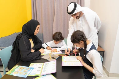 تنمية المهارات الثقافية والقدرات الفكرية للابناء، انشطة ترفيهية فنية ابداعية، لعبة تلوين الرسومات، زوجان عربيان خليجيان اماراتيان يرتديان الزي التقليدي يجلسان على الطاولة يقومان بتعليم ابنائهم، استغلال أوقات الفراغ بالأنشطة الترفيهية الممتعة