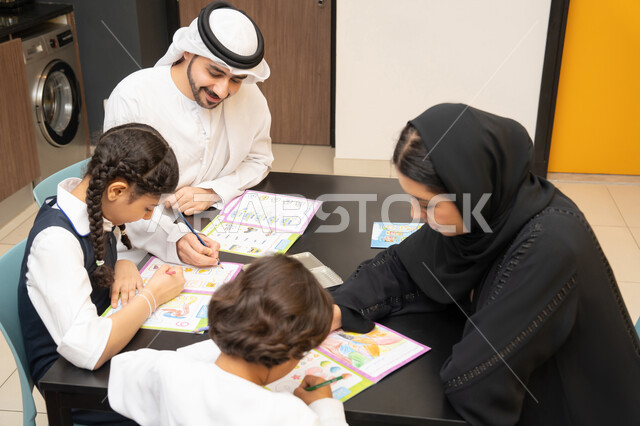 Caring for children's education, educational activities and developing children's learning abilities, supporting and encouraging children to solve lessons and homework, an Emirati Gulf Arab family wearing traditional dress sitting at a table and helping their children with education and reading