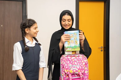 الاهتمام بتعليم بالأبناء ودعمهم، تعلم وتطوير الابناء، الاستعداد لبدء عام دراسي جديد، مفهوم التعلم في المدارس المتطورة في الامارات العربية، أم عربية خليجية اماراتية محجبة ترتدي عباءة سوداء تقوم بمساعدة ابنتها بتحضير الحقيبة المدرسية