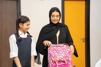 تعلم وتطوير الابناء، الاستعداد لبدء عام دراسي جديد، الاهتمام بتعليم بالأبناء ودعمهم ، مفهوم التعلم في المدارس المتطورة في الامارات العربية، أم عربية خليجية اماراتية محجبة ترتدي عباءة سوداء تقوم بمساعدة ابنتها بتحضير الحقيبة المدرسية